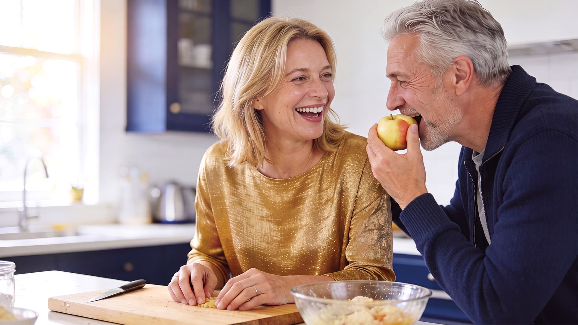 A smiling couple in their kitchen as the man confidently bites into an apple after dental implant treatment